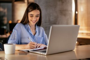 Woman writing on notepad, while using headphones and looking at laptop.