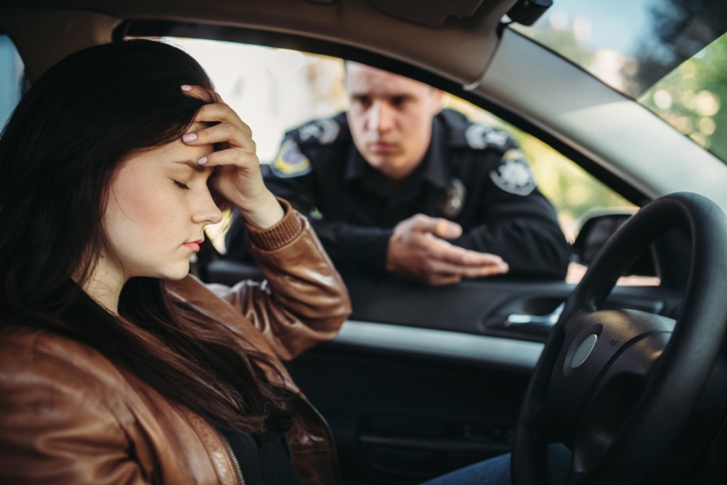 A woman in a car with her hand over her forehead, a stressed expression, and a policeman in the window.