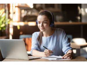 Woman studying on laptop while wearing head set and writing on notebook.