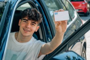Man holding license while sitting on car and smiling