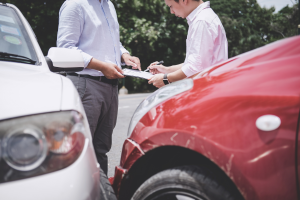 Two people stand near the front of a white car and a red car that appear to have been in a collision. One person is holding a clipboard while the other points to it with a pen, discussing or filling out paperwork.