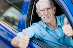 An elderly man with white hair, glasses, and a mustache leans out of the driver’s side window of a blue car, smiling broadly and giving two thumbs up.