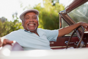An elderly man with a white hat and light blue shirt is sitting in the driver’s seat of a vintage convertible car. He is smiling broadly and looking toward the camera, with one arm resting on the car door and the other on the steering wheel. The background shows green trees and sunlight.