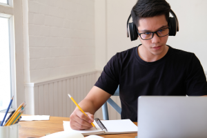 A young man wearing glasses, a black shirt, and headphones is sitting at a table with a laptop. He is writing in a notebook with a yellow pencil, focused on the laptop screen.