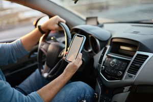 A person wearing a blue shirt is sitting in the driver’s seat of a car, holding the steering wheel with one hand and a smartphone in the other hand. The car's dashboard, controls, and center console are visible.