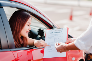 A woman with long brown hair is sitting in the driver’s seat of a red car, smiling and pointing at a clipboard with a car insurance form. A person outside the car, whose face is not visible, is holding the clipboard and a pen.