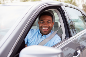 A man with short black hair, wearing a blue shirt and a seatbelt, smiles brightly while sitting in the driver's seat of a car. The car's window is rolled down, and the interior and exterior of the car are visible.