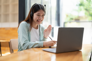 A young woman with long brown hair, wearing a headset and a light green cardigan, is sitting at a table with a laptop. She is smiling and waving at the screen while holding a pen in her other hand.