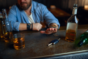 A man seated at a bar with multiple bottles of alcohol and a glass in front of him gestures to refuse the car keys on the counter.