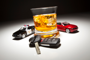 A glass of whiskey with ice, car keys, and toy police and sports cars placed together on a reflective surface, symbolizing the dangers of drinking and driving.
