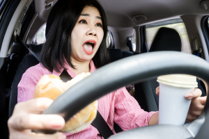A shocked woman holding a burger in one hand and a coffee cup in the other appears to be reacting to something while driving.