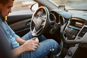A man sitting in the driver's seat is looking down at his phone, with the car dashboard visible in the background.