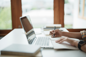 A pair of hands writing in a notebook beside a laptop on a tidy study desk with books.