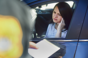 A woman sitting in her car looks worried while interacting with a police officer who is holding a notepad