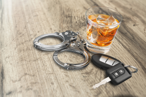 The image shows a pair of silver handcuffs next to a glass of whiskey over ice, with a car key resting on the table. The lighting is soft, and the image is arranged to highlight the juxtaposition of drinking and driving consequences.