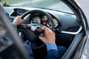A close-up shot of a person's hand holding a bottle of beer while driving a car. The driver is gripping the steering wheel with one hand while holding the bottle in the other. The image captures the action of drinking while driving.