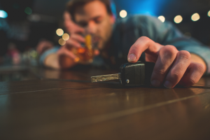 A man is sitting at a table in a bar or restaurant. He is reaching for his car keys while holding a glass of alcoholic drink in the other hand. His facial expression suggests he might be intoxicated.