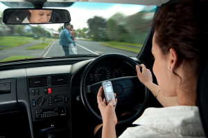 A woman is driving a car while looking down at her mobile phone. The steering wheel is visible along with part of the dashboard as she holds the phone in her left hand.