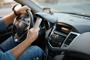 A close-up shot of a driver's hands on the steering wheel and holding a smartphone. The individual appears to be texting while driving, with the road visible ahead through the windshield.