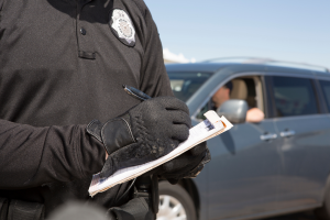 A police officer is writing a ticket while standing next to a car. The officer is wearing a black shirt and gloves, and a pen is in his hand as he prepares to issue the ticket. The car’s driver is inside, but not visible in the frame.