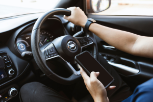 A person is seen driving a car while holding a smartphone in their left hand, possibly texting or checking it. The image focuses on the driver's hands on the steering wheel and the phone.