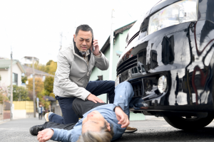 Man kneeling next to man that is laying on street, car appears to have ran over him.