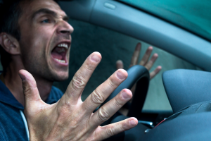 A man is driving a car, shouting with an open mouth and raised hands off the steering wheel, suggesting frustration or anger. The interior of the car is visible, with a blurred outdoor background.