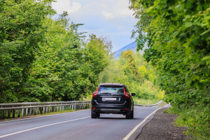 A black SUV is seen from behind, driving down a winding road flanked by dense green trees and a guardrail. The background includes distant hills or mountains under a partly cloudy sky.