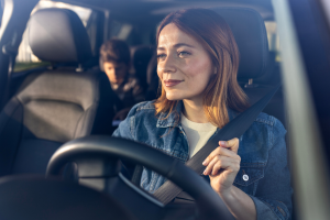 A smiling woman wearing a seatbelt is driving a car, with a child visible in the backseat.