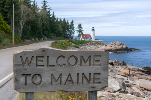 A wooden sign reading "WELCOME TO MAINE" is prominently displayed along a roadside. In the background, a lighthouse with a red roof sits on a rocky coastline beside the ocean. The scene includes a road bordered by trees and greenery under a partly cloudy sky.