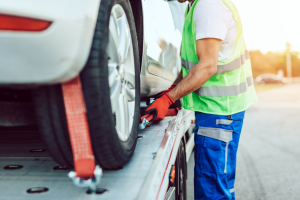 A tow truck operator in a reflective vest and gloves securing a vehicle's tire with a strap on a flatbed truck.