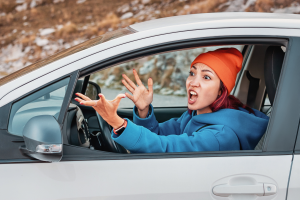 person is driving a car, with one hand raised in a gesture and the other on the steering wheel. The driver is wearing a blue jacket, and the scene appears to be at night or in low light, with a focused view inside the car.
