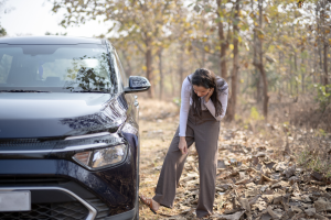 A woman stands beside a black car on a rocky, forested path, appearing to lean on the vehicle with a contemplative expression. She is dressed in a white top and brown pants, with a blurred natural background of trees and dry leaves.