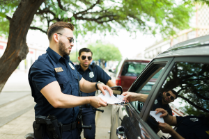 Two police officers handing documents to a driver at a car window, with a tree-lined street in the background.