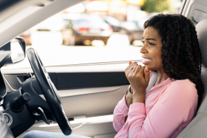 A person inside a car with a worried expression, hands near their mouth, wearing a pink shirt.