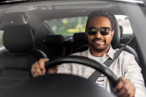 A man wearing sunglasses, smiling while driving a car. Sunlight is shining through the windows, and he appears relaxed and focused on the road.