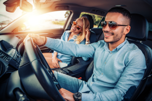 A man and a woman sitting in a car together, both wearing sunglasses and smiling. The sun is shining through the windshield, creating a bright, cheerful atmosphere.