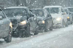 A line of cars driving closely together on a snow-covered road during heavy snowfall. Headlights are on, and snowflakes fill the air, reducing visibility and indicating winter driving conditions.