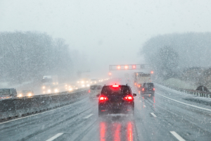 A highway scene with cars driving in snowy, low-visibility conditions. Red tail lights and road signs are visible through the falling snow.