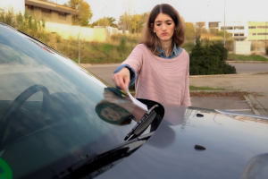 A woman picking up a parking ticket or notice under her car’s windshield wiper, looking displeased. The setting appears to be a parking area on a clear day.