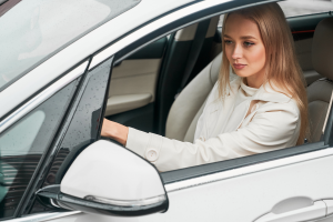 A woman sitting behind the wheel of a white car, looking focused ahead. She is wearing a light-colored coat, and raindrops are visible on the car’s exterior.