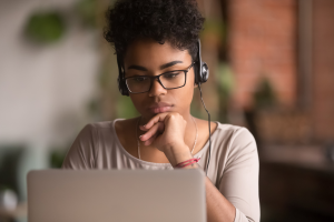 A young woman wearing glasses and a headset, concentrating while looking at a laptop screen.