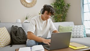 Young man relaxing, sitting on the sofa in his living room, immersed in studying online with his laptop, listening to musiC.