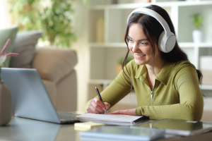 Woman using laptop to study. She us wearing headphones and has a notebook and pen.